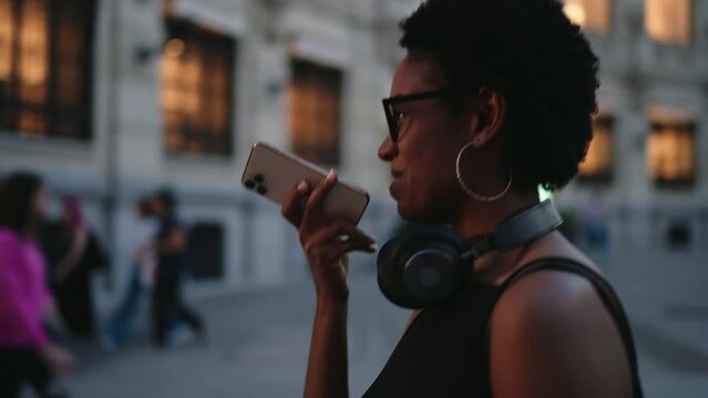 Radiant young woman with stylish glasses and over-ear headphones engaged in lively conversation on smartphone, with the city's early evening ambiance as backdrop. Black female enjoy audio message app