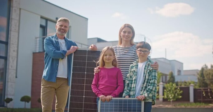 Happy family looking at camera with solar panel. Parents and children smiling at background of house exterior. Mother and father instilling in kids love of planet. Eco awareness concept.