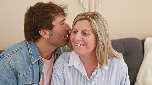Caucasian mother and son share intimate secret whilst casually lounging on the living room sofa together at their cozy home, reflecting joy, confidence and a relaxed lifestyle.