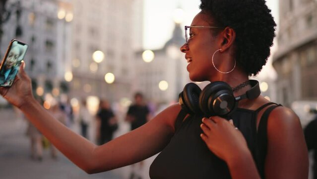 Smiling African-American female engages in vibrant video call, her phone's front camera catching golden city hues at dusk. Happy black woman enjoy online communication with her family despite distance