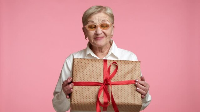 Well-groomed old lady in spectacles swoon with happiness, opening mouth with excitement, pressing present in golden paper with red ribbon, pink background. High quality 4k footage
