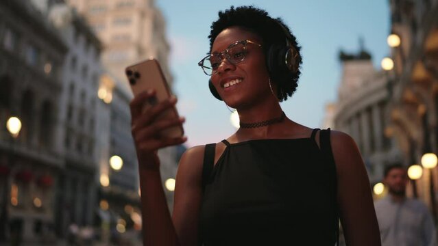 Smiling female blogger scrolling social media while walk with headphones in evening city. Cheerful young black woman enjoy listening to music,reading text messages on her smartphone device.Slow motion
