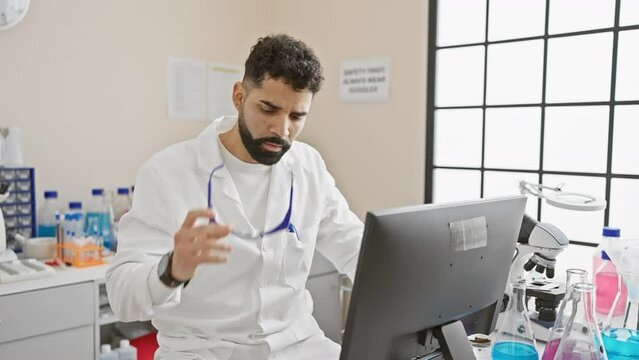 A young hispanic man with a beard wearing lab coat and safety glasses works at a computer in a laboratory setting.