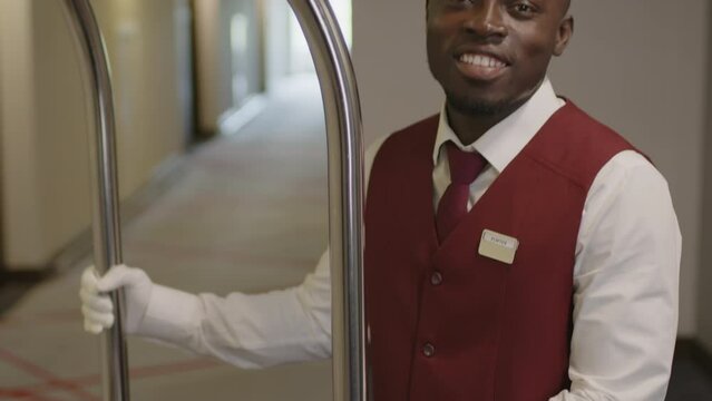 Tilt up portrait shot of young Black bellhop in elegant uniform and white gloves happily smiling on camera while posing with luggage cart in hotel hallwayTilt up portrait shot of young Black bellhop i