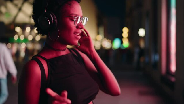 Joyful woman dancing on night neons street, headphones on, feeling rhythm of favorite music.Slow motion. Happy and carefree African American female moves her body with relaxed song beats in earphones