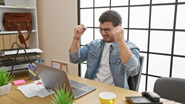 An overjoyed man in an office crossing fingers for good luck near a laptop.