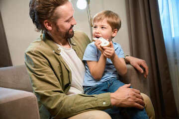 Little boy eating gingerbread while sitting together with father in hotel