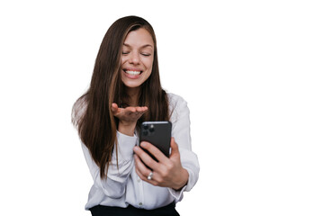 Fototapeta premium Cheerful Hispanic young woman holds phone sends air kiss during video call against transparent background dressed in white shirt. Pretty businesswoman grateful to husband. Happy woman