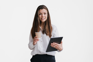 Fototapeta premium Young purposeful brunette businesswoman in white shirt and black pants holding tablet and cup of coffee looks at camera toothy smiles against transparent background. Smart student isolated