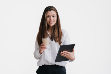 Young purposeful brunette businesswoman in white shirt and black pants holding tablet and cup of coffee looks at camera toothy smiles against transparent background. Smart student isolated