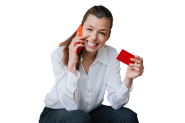 Successful young woman in shirt and black pants sitting against transparent background talking by phone showing credit card looking at camera. Business people and wealthy life. Online purchase