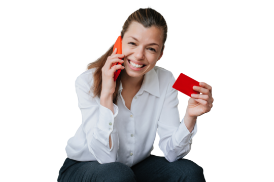 Successful young woman in shirt and black pants sitting against transparent background talking by phone showing credit card looking at camera. Business people and wealthy life. Online purchase