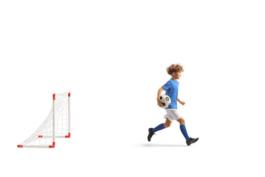 Full length profile shot of a boy in a football kit running and carrying a ball in front of a goal