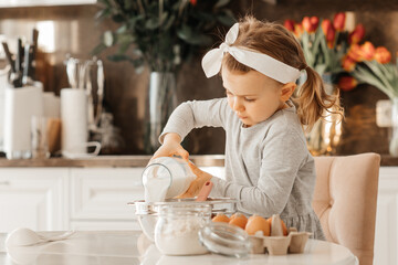 Funny child girl cooking cake for Easter party in white sunny kitchen with spring flowers. Home...