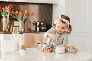 happy excited kid girl baking cake for Easter emotional child cook dessert present for mother day in white sunny kitchen Home bakery. Bottle of milk, eggs, flour are on the table