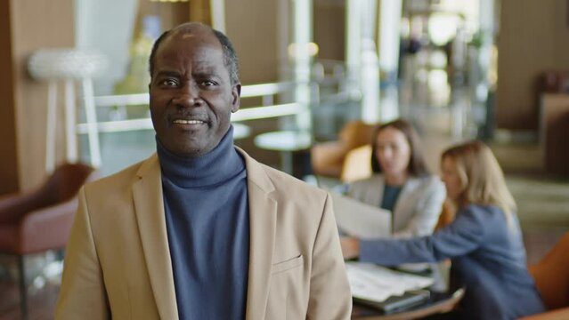 Medium shot of middle aged Black businessman in smart casual outfit standing in hotel restaurant and posing for camera with smile, female colleagues having meeting in background