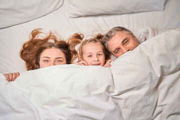 Mother, father and daughter covering face with blanket on bed in hotel room
