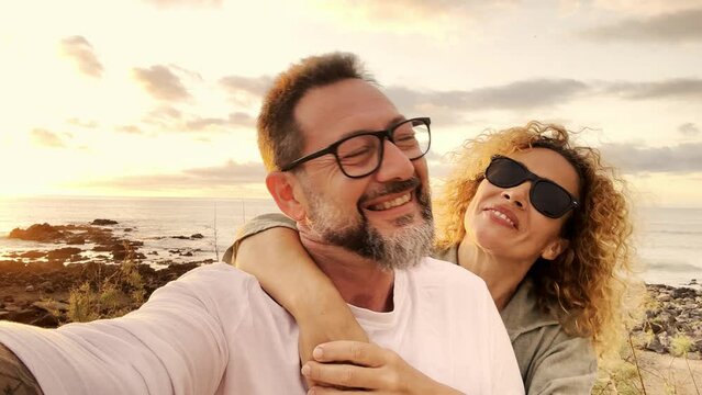 Happy couple of adult tourist enjoy summer holiday vacation kissing and having fun together at the beach against a golden sunset on the ocean. Man and woman do selfie video in outdoor leisure holiday