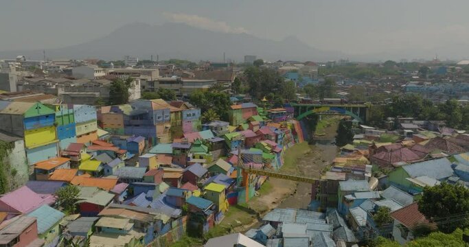 Aerial Shot Of Colorful Residential Houses In Village By Mountain, Drone Flying Forward Over Canal On Sunny Day - Java, Indonesia