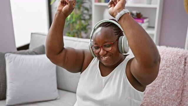 Cheerful plus size african american woman, with braided hair and glasses, confidently dancing and enjoying music while sitting, relaxed on her living room sofa at home.