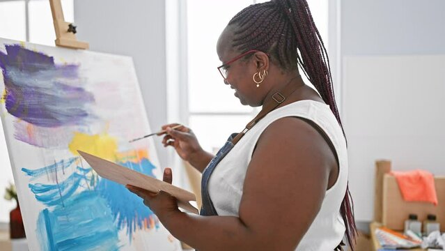 Confident, smiling african american woman artist joyfully drawing in art studio with her brush and canvas, embracing creativity.