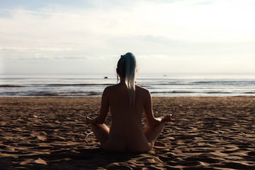 Rear view of silhouette of bare nudist lady sit asana yoga lotus pose on nudist wild beach. From...