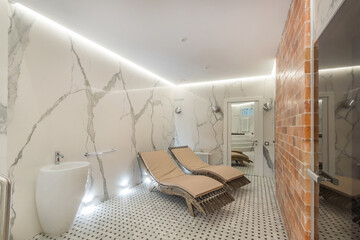 A luxurious bright bathroom with a brick wall, two sun beds and a white original sink.