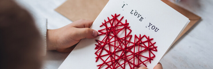 Little kid preparing diy handmade cute post card for Mother's Day with a message I love you mom. Red hear embroidering, printing. Concept of simple presents, family values, hobby, leisure activity
