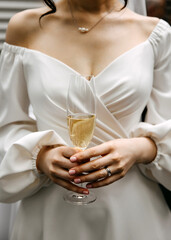 A bride in an elegant off-the-shoulder white dress holding a champagne flute, wearing a diamond ring.