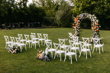 Outdoor wedding setup with floral arch. Wedding aisle with white wooden chairs.