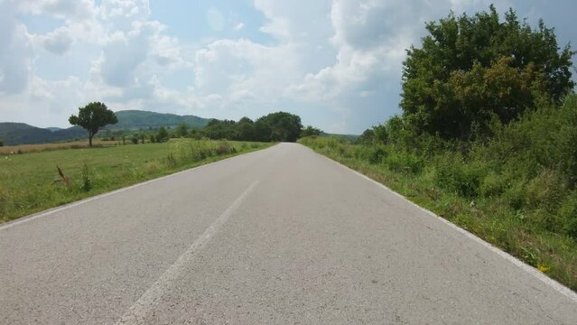 A very fast car or motorcycle ride on a bumpy winding country road that passes between agricultural fields and trees. A mountain peak visible in the distance. Blue sky with lots of clouds, summer time