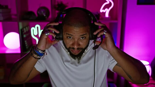 A bearded man with headphones in a neon-lit gaming room engages with technology at night