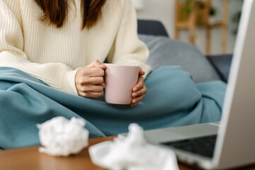 Close-up view of young unrecognizable sick woman drinking hot drink while using laptop at home