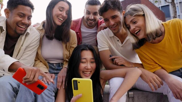 Group young people laughing watching something on phone. Multiracial friends sitting outdoor using mobile together. Concept of friendship social networks and technology addiction in new generations.