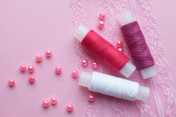 Fototapeta na wymiar Cotton red and white threads in spools and pink beads lie on pink lace. View from above