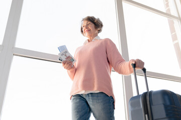 Low view european old elderly senior woman traveler with suitcase bags luggage baggage holding passport and airline tickets walking through airport train station waiting for flight, looking for gate