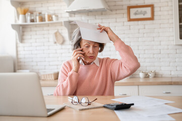 Angry confused stressed old elderly senior woman grandmother having trouble failure problem issue with documents, papers, making mistake error, working remotely and talking on cellphone at home