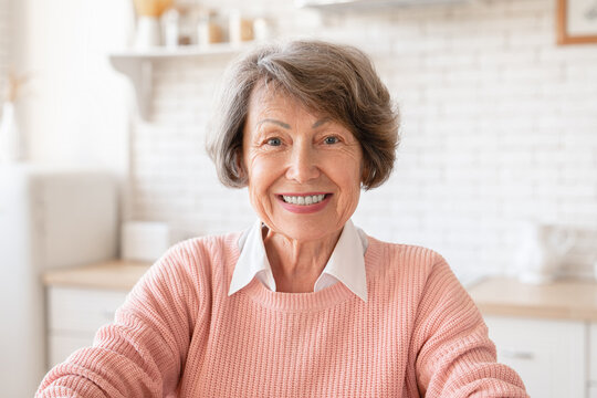 Closeup cropped portrait of senior old elderly caucasian woman grandmother housewife looking at camera with toothy smile at home kitchen. Videocall concept