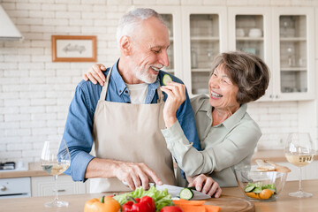 Cheerful old senior wife treating her husband while helping him cook prepare dinner salad lunch at home kitchen. Love and care, togetherness concept