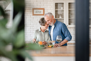 Love and care concept. Quality time and acts of service - love language. Senior old couple grandparents hugging cuddling helping prepare food cook romantic dinner and drinking wine at home kitchen