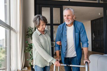 Rehabilitation after traumas injuries. Old elderly senior woman caretaker nurse wife supporting helping disabled handicapped man patient husband to move with walking frame