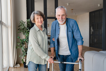 Happy senior couple looking at camera. Disabled incapacitated handicapped old elderly husband using walker walking frame for moving while wife helping him - obrazy, fototapety, plakaty