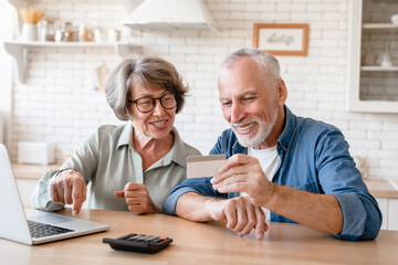 Cheerful caucasian senior old couple grandparents using credit card for paying domestic bills, receiving money pension, e-banking, e-commerce at home on laptop