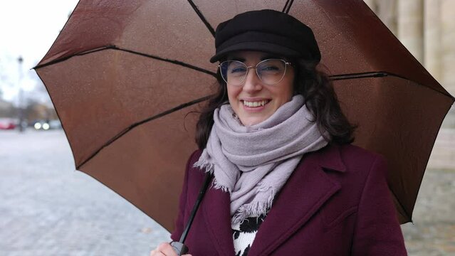 Cheerful portrait of young adult woman in warm clothes holding an umbrella while looking at camera on a rainy day at city street.
