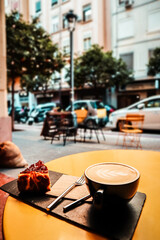 Cup of art latte or cappuccino coffee on a yellow round table on a cafe terrace. Breakfast hot drink beverage and sweet pie. Autumnal drink in a mug.