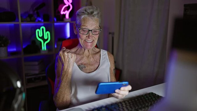 Epic win! grey-haired senior woman streamer, gaming guru, celebrates big win during late-night video game stream in a dark gaming room, smartphone in hand!