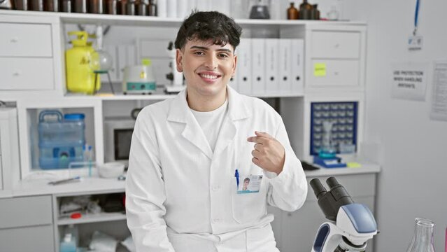 Confident young male scientist, cheerfully pointing with hand and finger, flashes a joyful, hopeful smile while sitting in lab