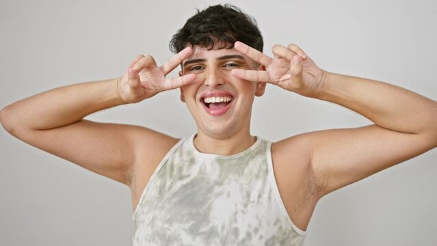 Cheerful young man wearing sleeveless t-shirt, smiling while making peace symbol on face, proudly showing victory gesture on an isolated white background.