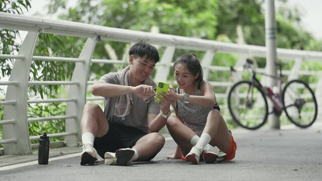 happy young asian couple sitting on ground outdoors looking at cellphone photos together while taking a break from exercise