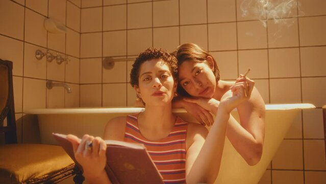 Young beautiful lesbian couple smoking a cigarette, holding a book and posing for camera in tiled bathroom with warm yellow light. Video portrait